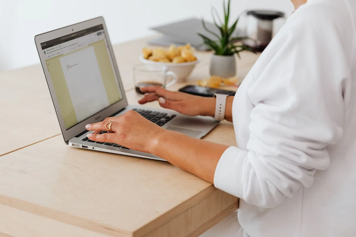 Person typing on a laptop at a wooden desk with a coffee cup and a small plant nearby
