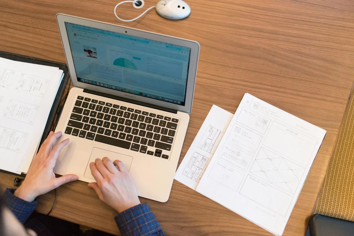 Person working on a laptop with documents and design files spread across a desk