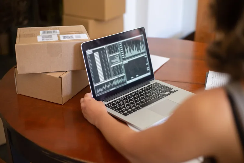 Small business owner reviewing online listings from a laptop with shipping materials nearby