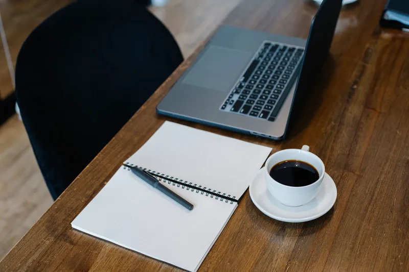 Laptop and notebook on a wooden table beside a cup of coffee and a pen