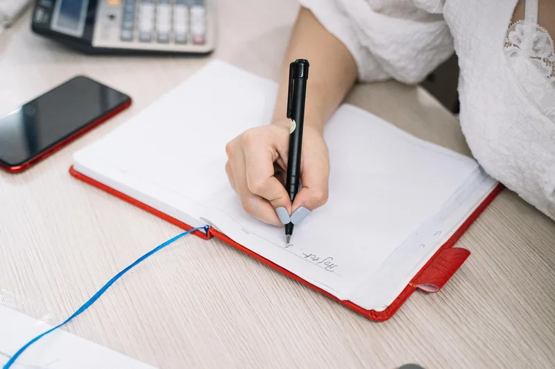 Adult hand writing notes in a notebook with a phone and calculator on the desk