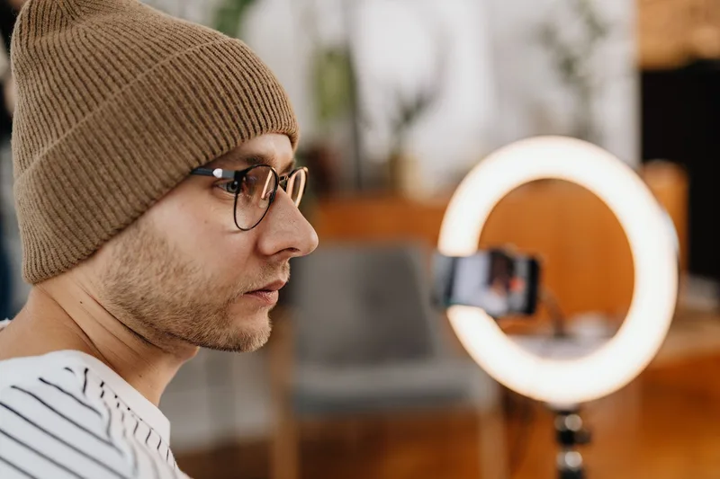 Man in glasses and a knitted cap recording a video at home with a ring light and smartphone